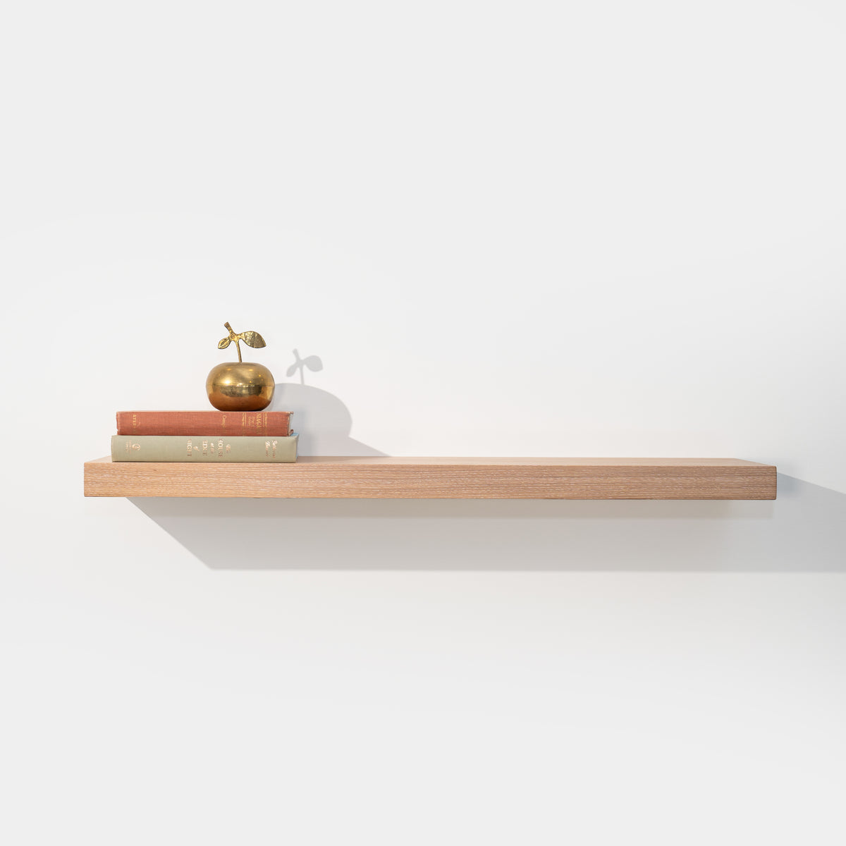 Solid white oak floating shelf with books and a brass apple on a white wall